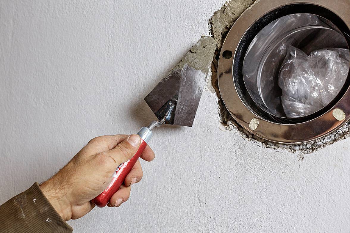A hand is applying mortar around a circular ventilation pipe on a white wall using a plastering trowel.