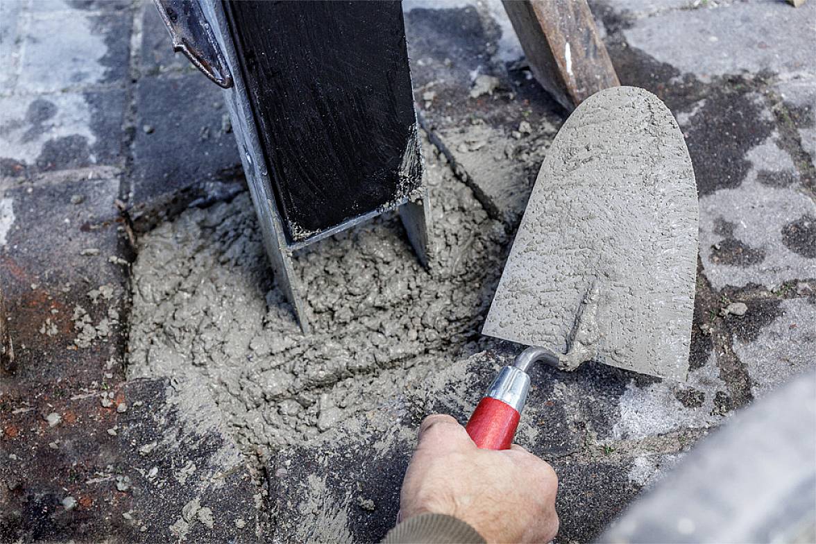 A hand holds a trowel with fresh concrete being poured around a metal post foundation on a paved floor.