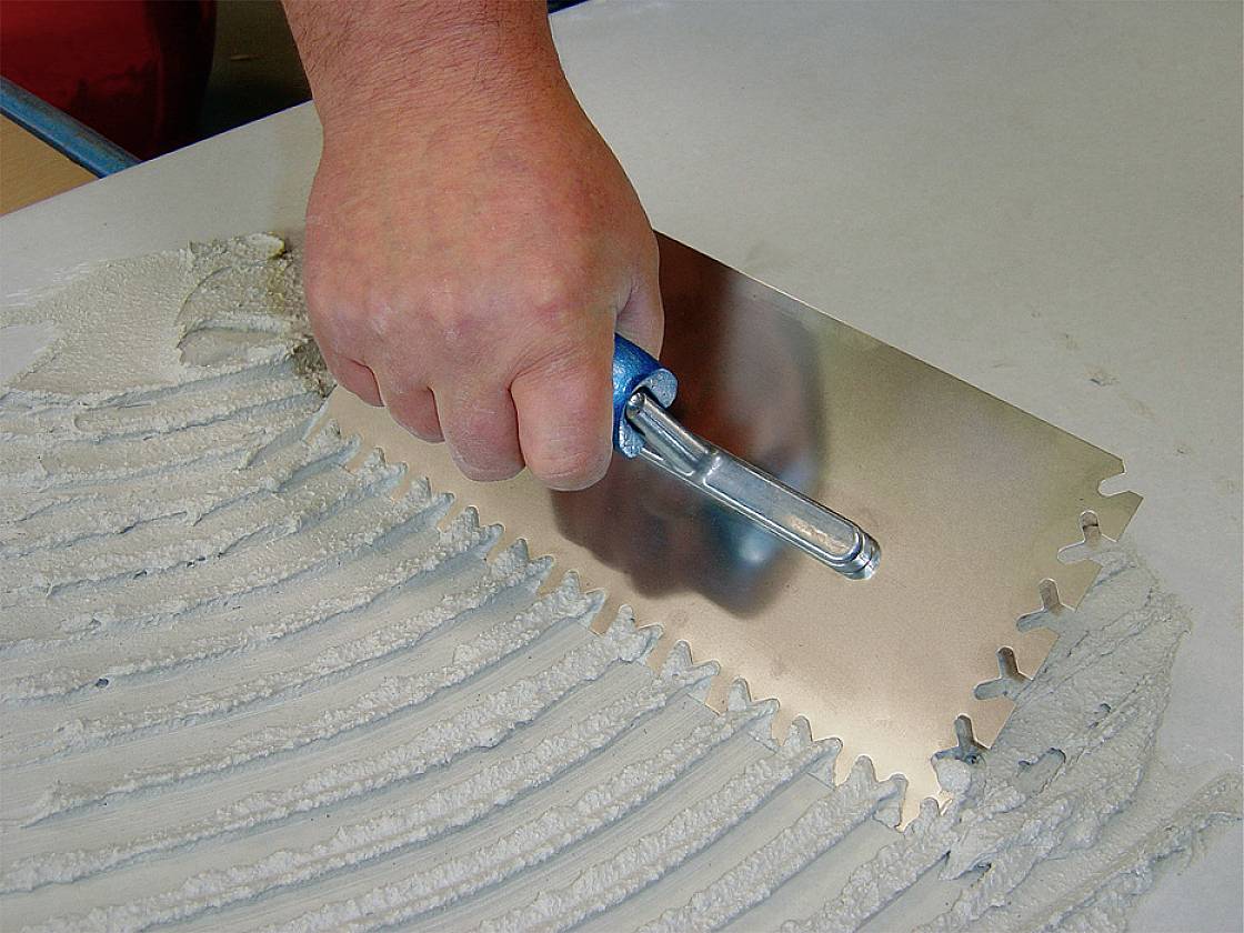 A hand holds a serrated trowel and spreads adhesive evenly across a surface.