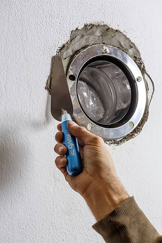 A hand is applying plaster around a circular ventilation pipe in the wall. The tool has a blue handle. Repair or construction work.