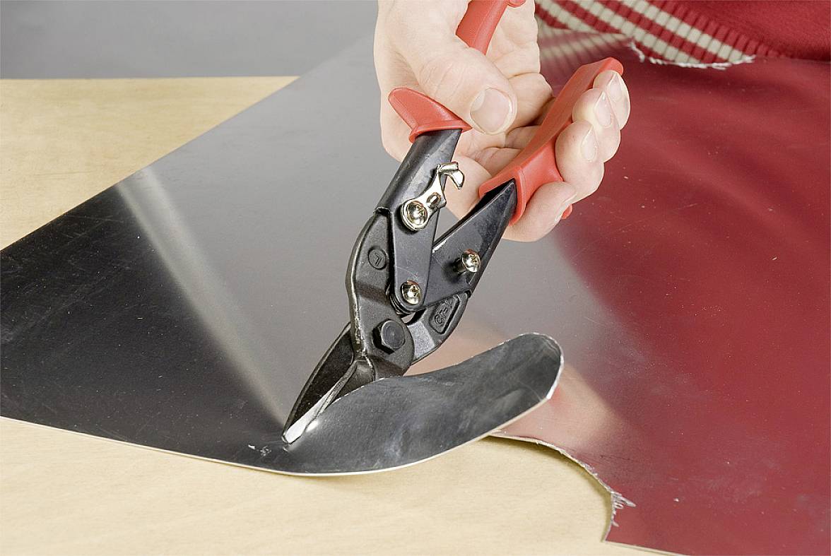A hand is cutting a piece of metal with a red tin snips on a wooden surface.