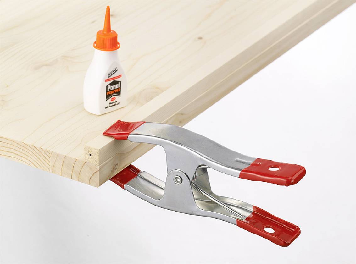 'Thin wooden board secured at the edge with a red clamp, next to a small bottle of wood glue on a white table surface.'