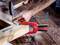 A person is hammering a nail into a piece of wood that is secured with a red clamp. An angle gauge is lying next to it on the table.