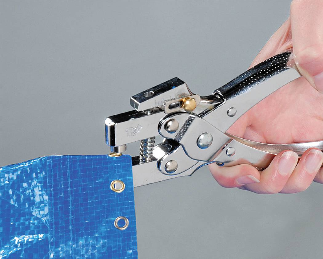 A hand is holding a metal pliers inserting eyelets into a blue tarpaulin.