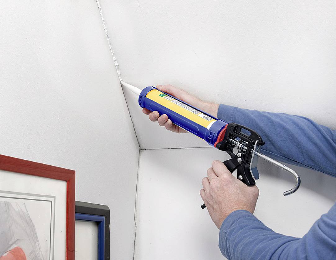 A person is using a cartridge gun to apply a white sealant to cracks in a wall; frame in the foreground.