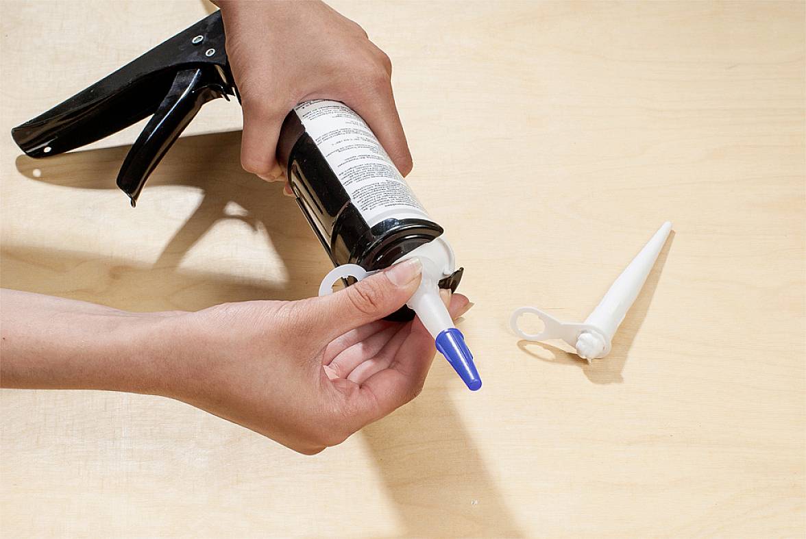 A person is using a cartridge gun to apply sealant. A nozzle is lying beside it on the table surface.