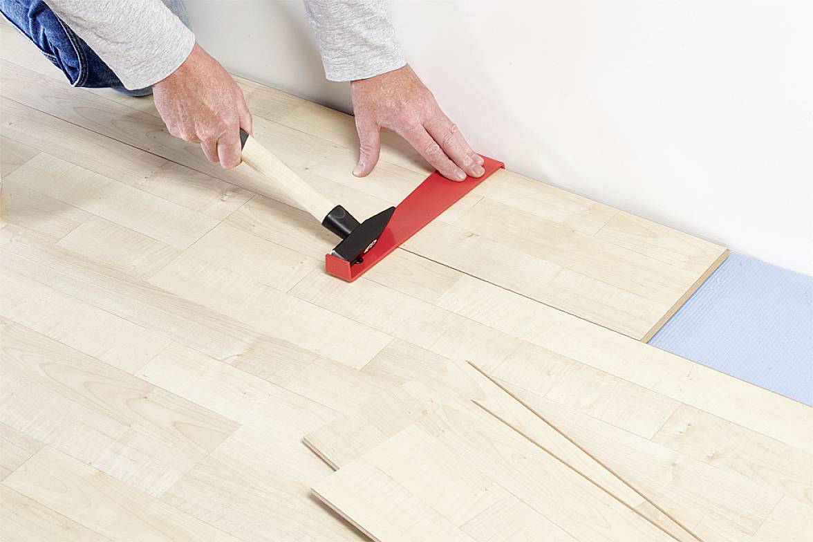 A person is installing a wooden floor, using a hammer and a red installation tool to secure boards precisely against the wall.