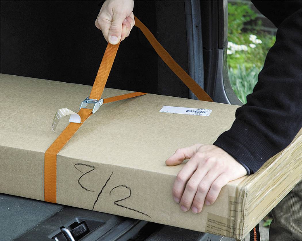 A person is securing a rectangular package in a car boot with an orange ratchet strap.
