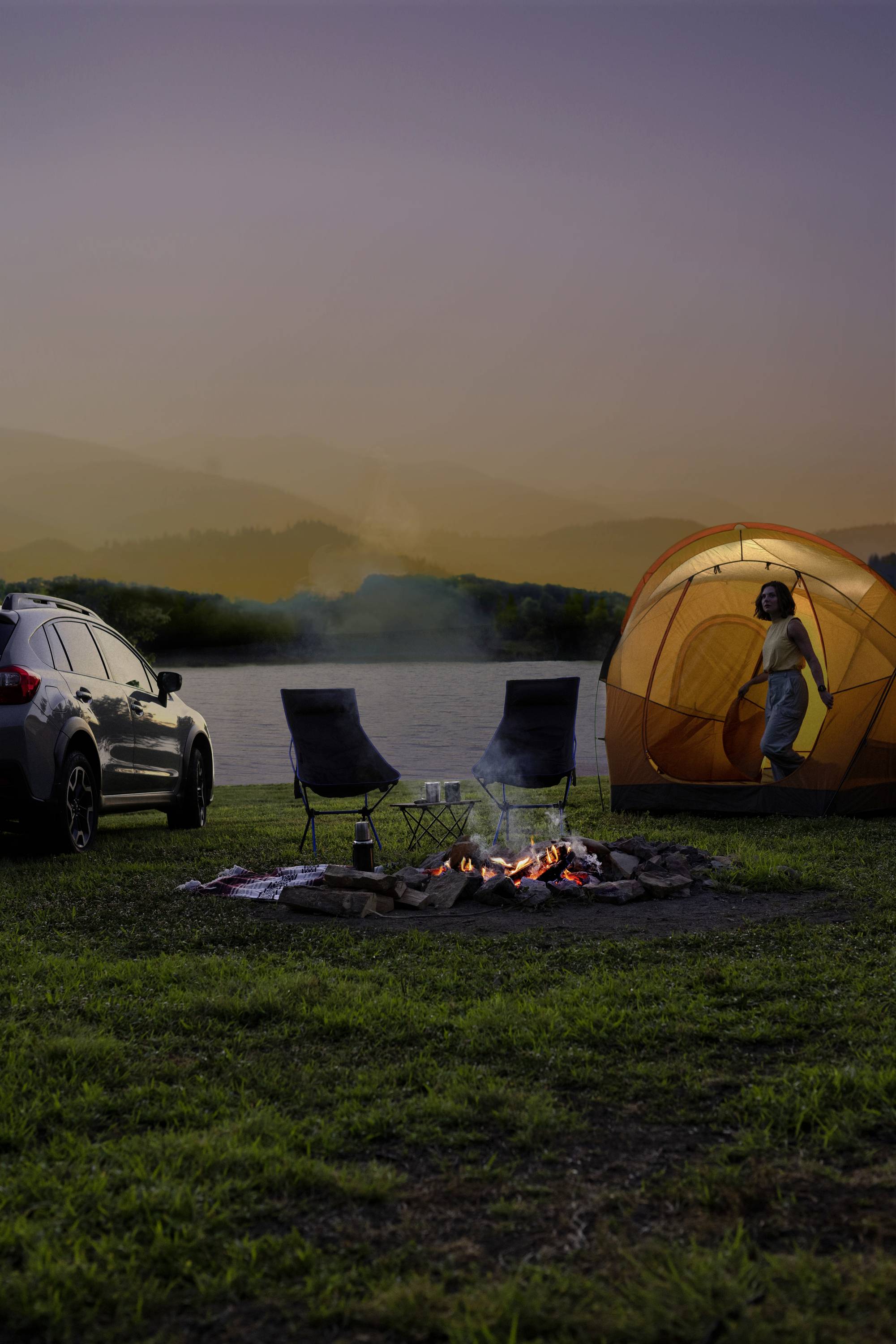 Car beside tent on lakeside at sunset. Two chairs and campfire in foreground, person looking out from tent.