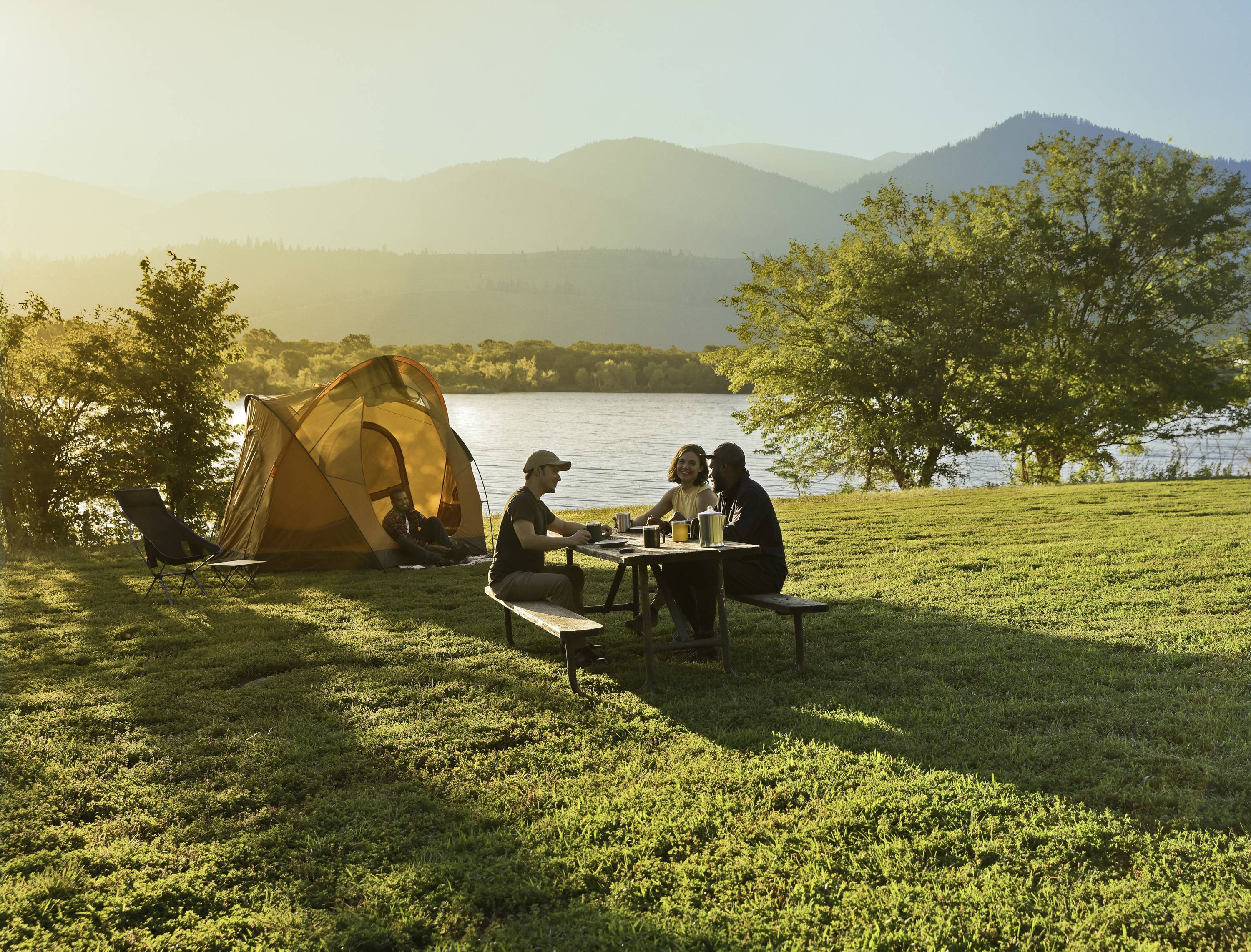 'Three people are sitting at a picnic table beside a tent by the lake. The sun is setting, and mountains can be seen in the background.'