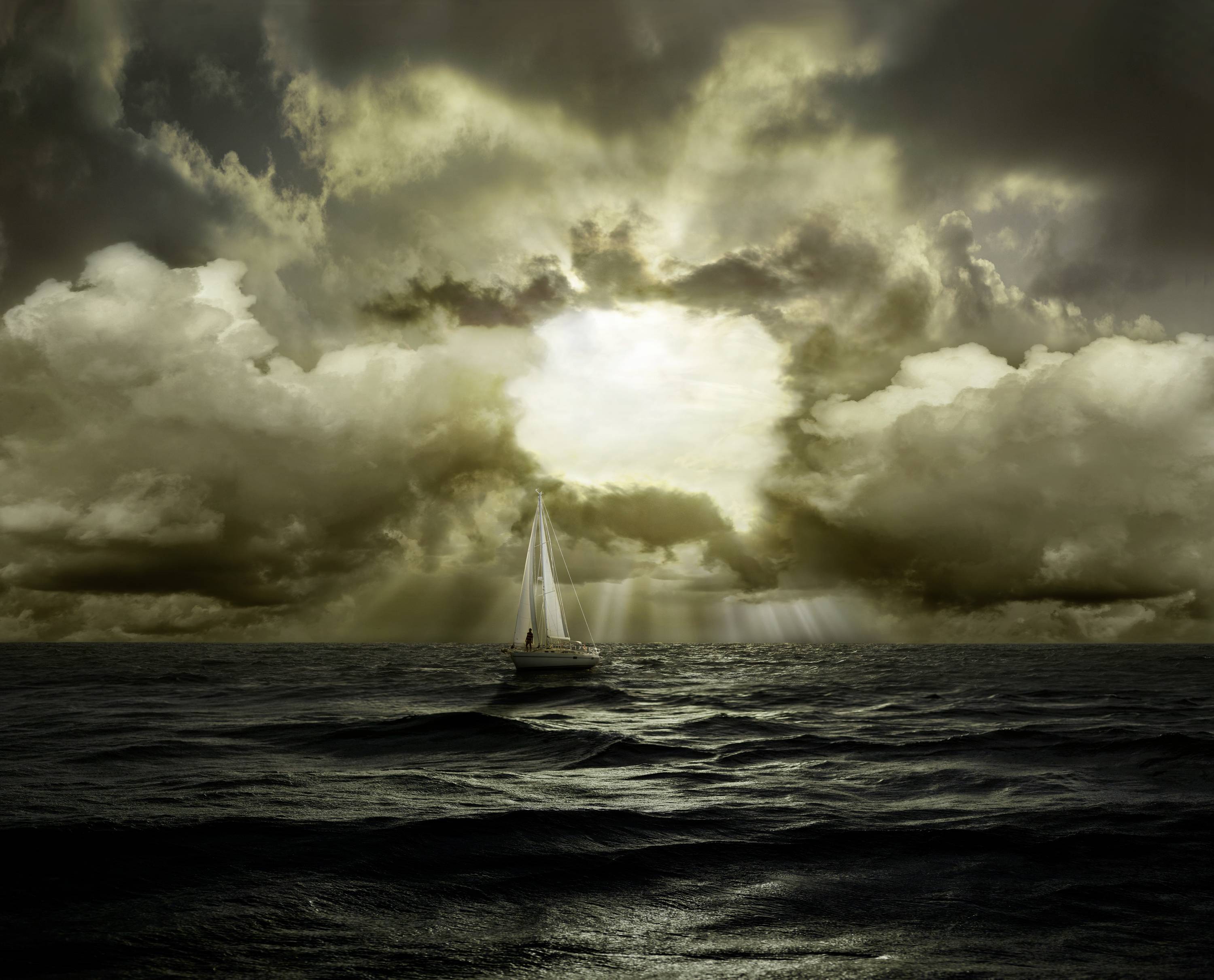 A sailing boat glides across a stormy sea beneath a dramatic, cloud-laden sky, pierced by rays of light.