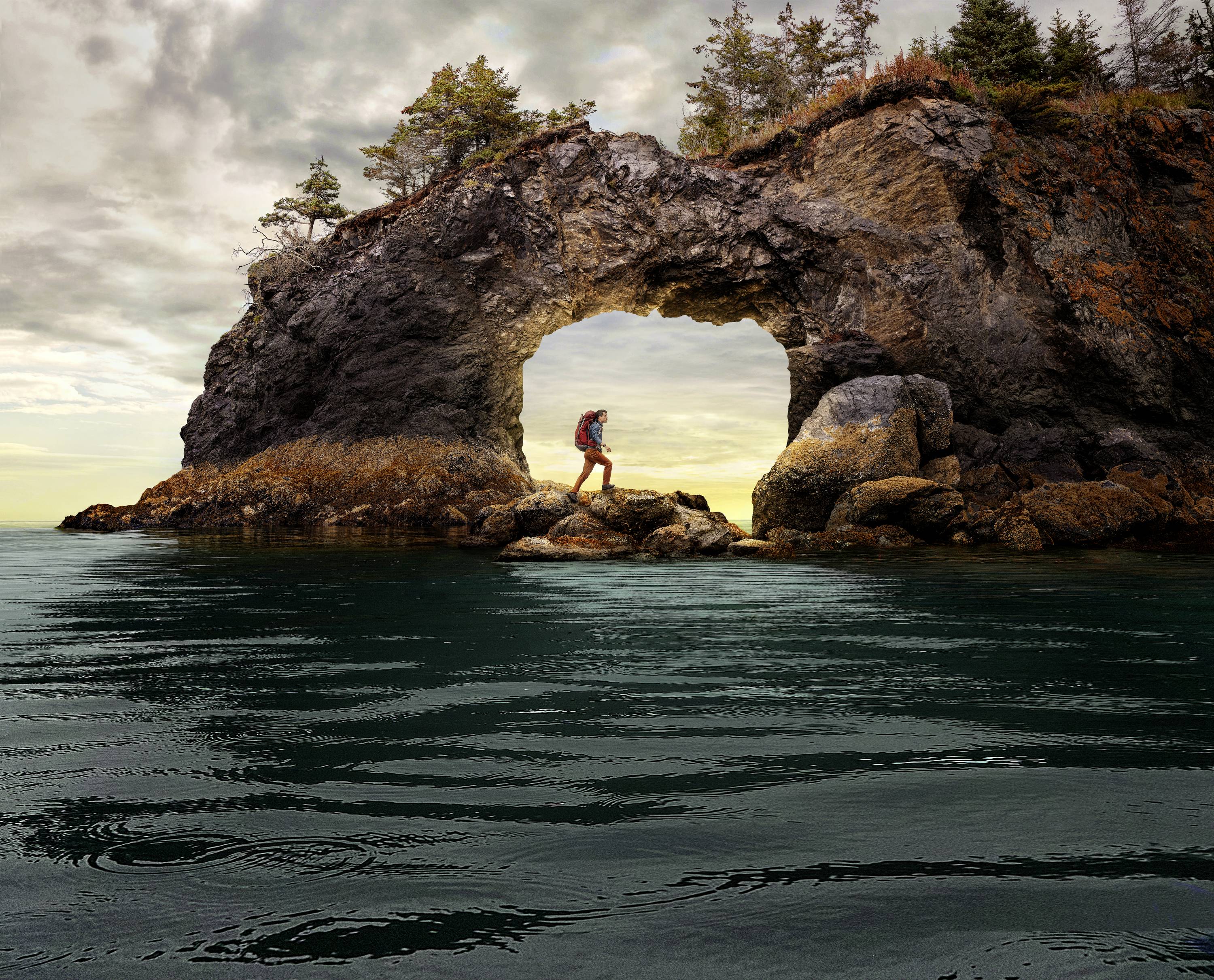 A person with a rucksack stands on rocks beneath a natural rock arch by the sea, surrounded by water and trees.