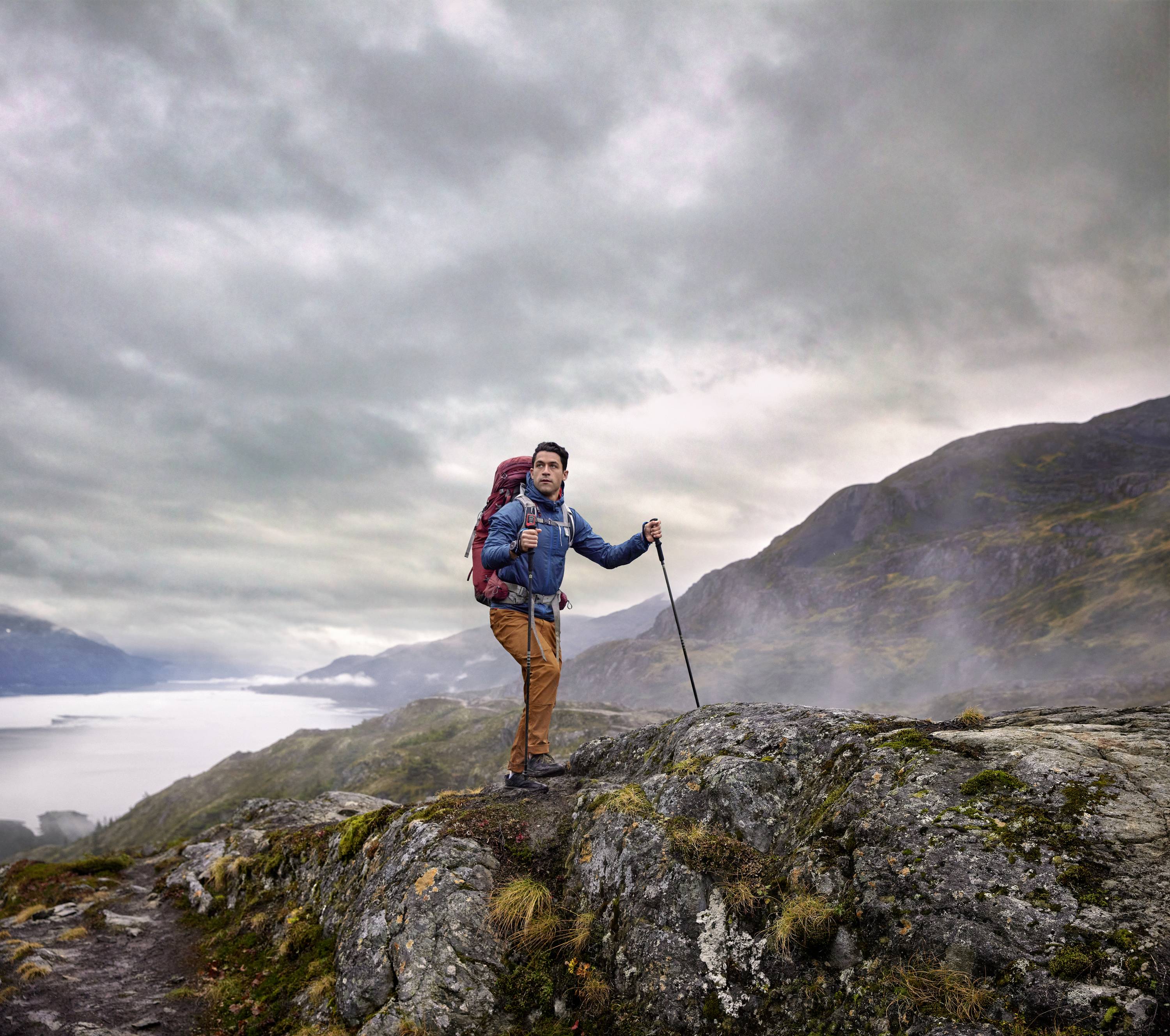 A hiker with a rucksack and walking stick stands on a rocky path. In the background, misty mountains and a body of water can be seen.