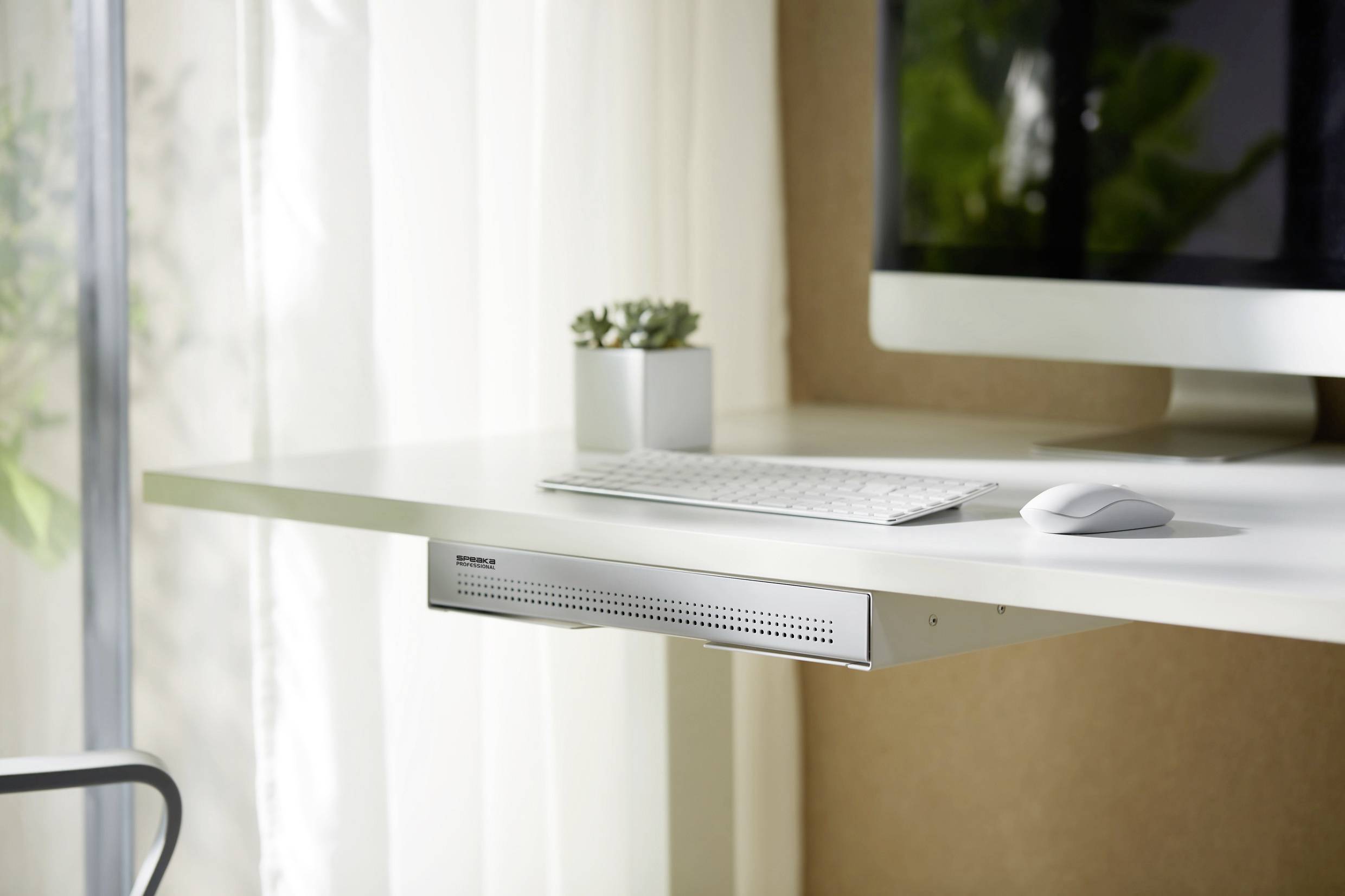 A modern, white desk with a keyboard and mouse, with a monitor positioned above. A small plant sits beside it. Light curtains in the background.