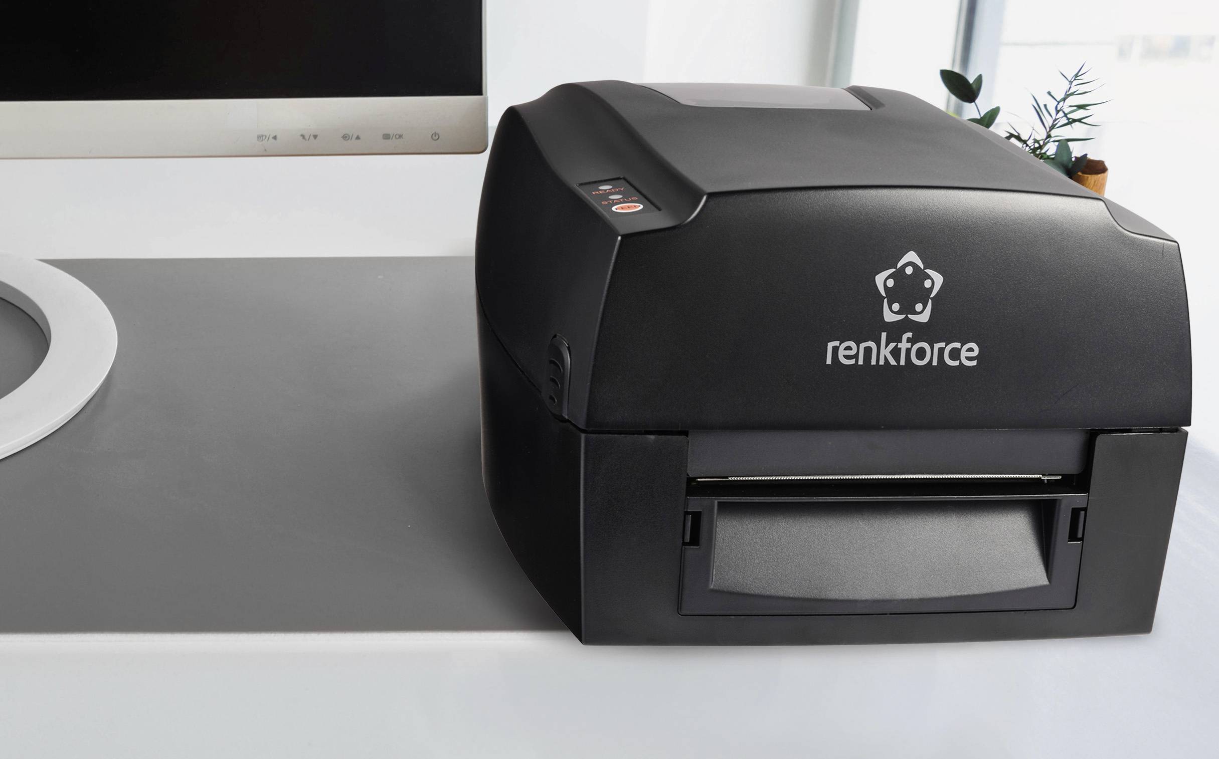 A black Renkforce label printer sits on a grey table, surrounded by electronic devices and plants in the background.