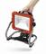 A hand is holding a portable, red and black LED work light with a sturdy stand, suitable for use on construction sites.