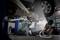 A mechanic is repairing a car that is raised on jack stands in a workshop. A lamp illuminates the work area. Tyres are visible.