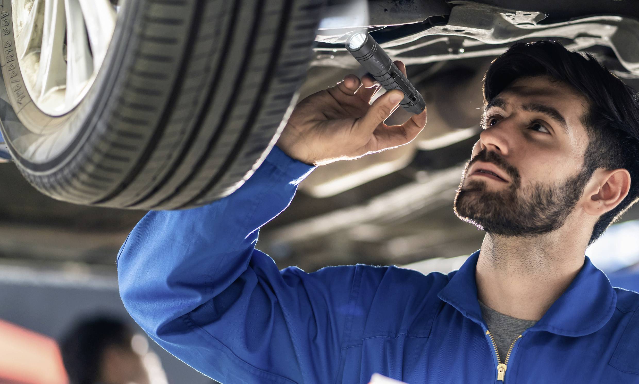 A man in blue workwear is inspecting the underside of a car with a torch in a garage.