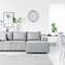 Grey sofa in a bright living room with wall shelves, plants, and a round clock. Modern, minimalist interior design style.