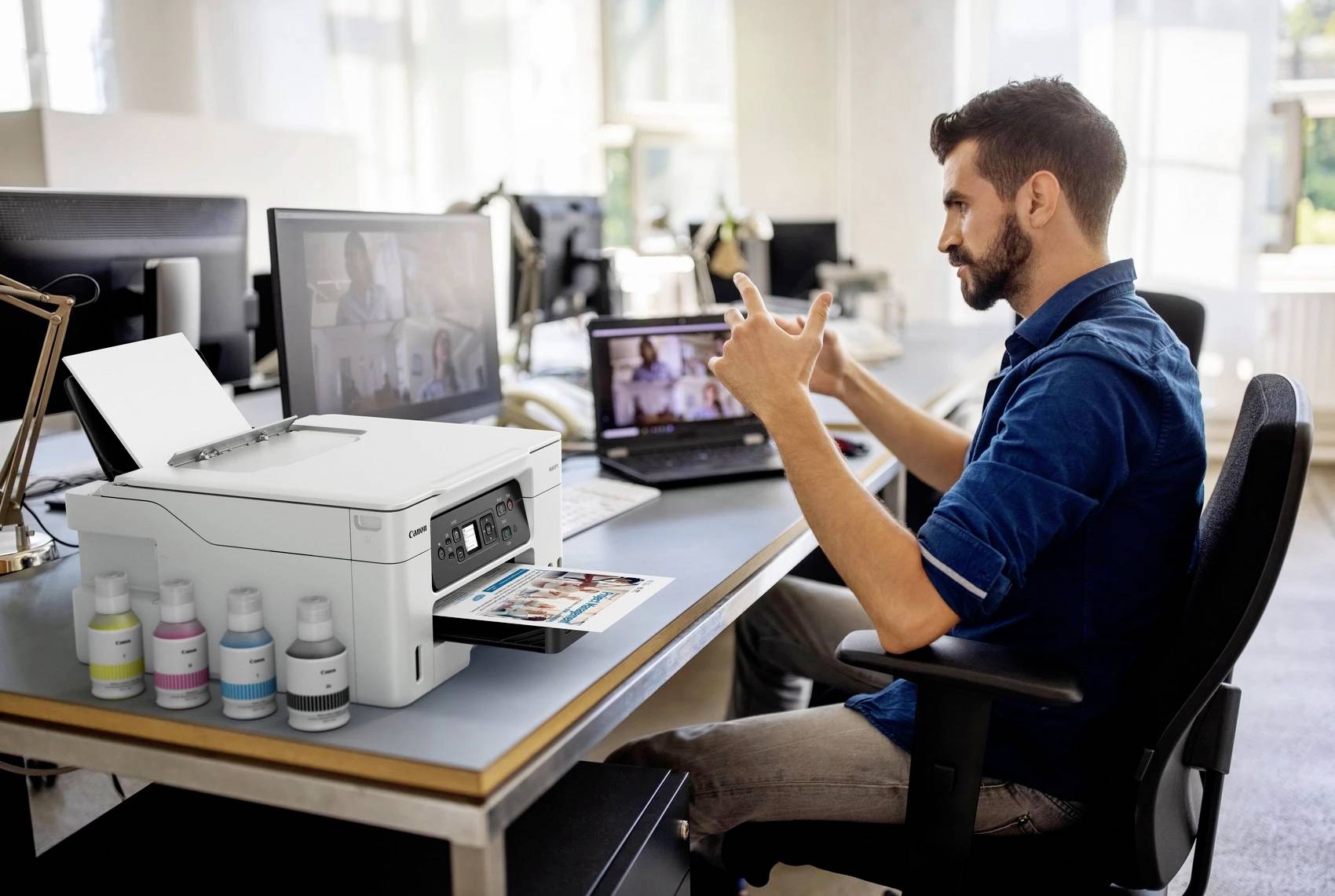 A man is sitting in an office at a desk, operating a laptop and looking at the screen. A printer is standing next to him.