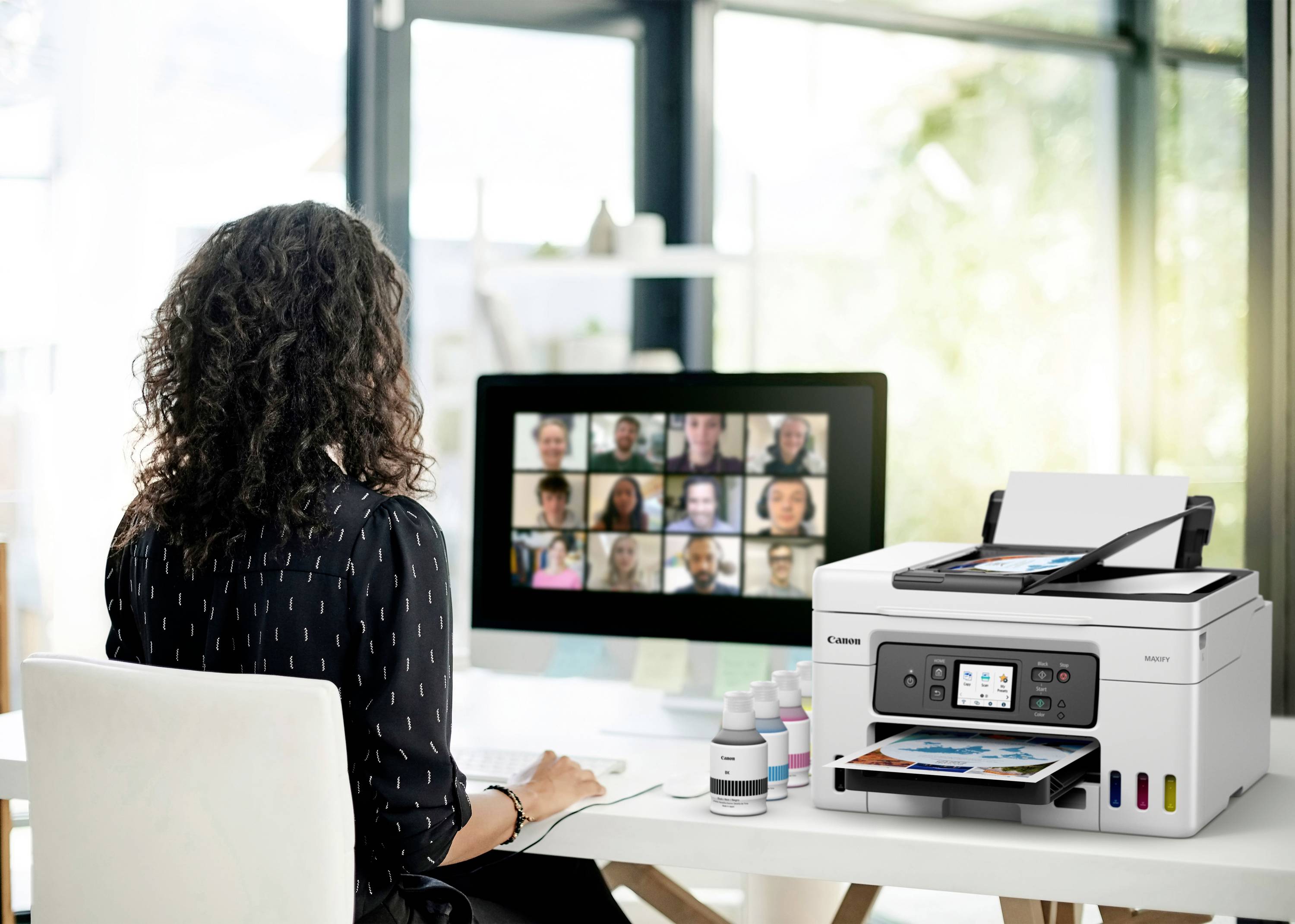A person is sitting at a desk and using a computer for a video conference. A printer with printed photos stands beside them.
