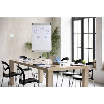 Meeting room with a table surrounded by chairs. A flipchart displays diagrams. Windows in the background allow light to enter.
