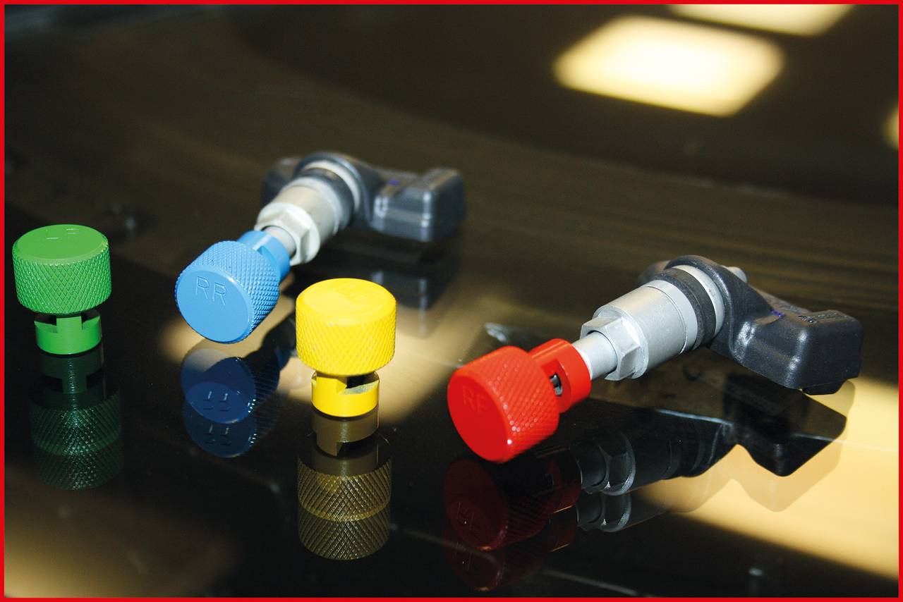 Four screw valves on a table, each in a different colour: green, blue, yellow, and red. Each valve has a metallic handle.