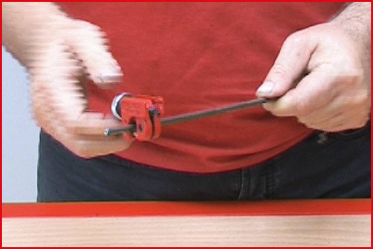 A person is cutting a thin metal pipe with a red pipe cutter on a table.