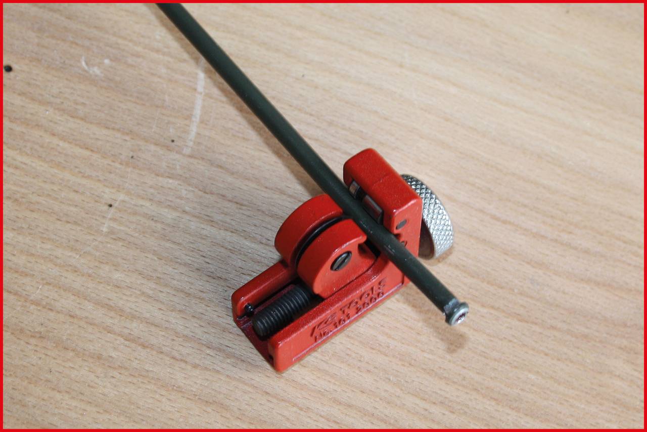 A small, red pipe cutter is resting on a wooden table, gripping a thin metal rod, ready to cut.
