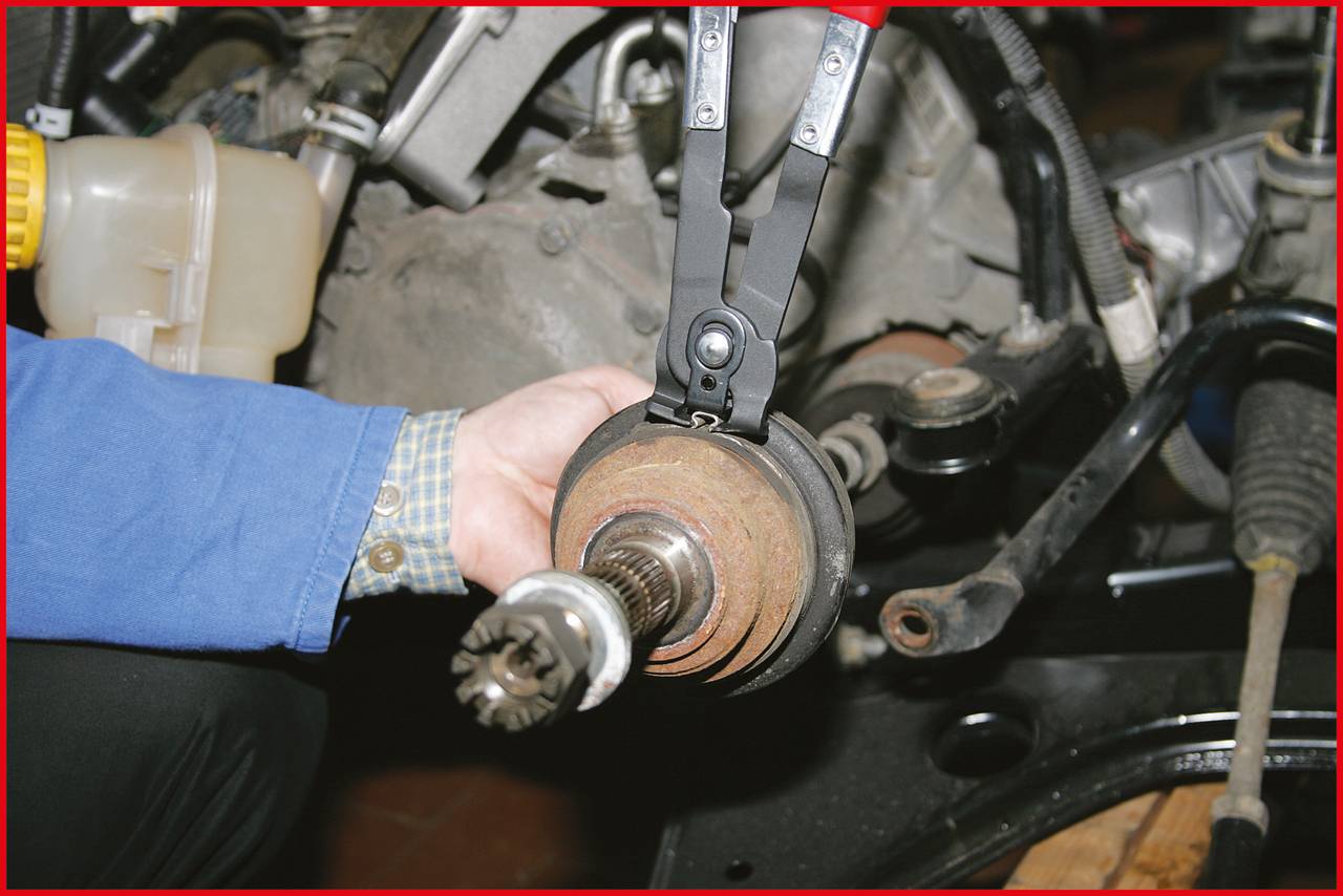 Someone is holding a replaced drive shaft next to a vehicle engine. In the background, engine parts and hoses can be seen.