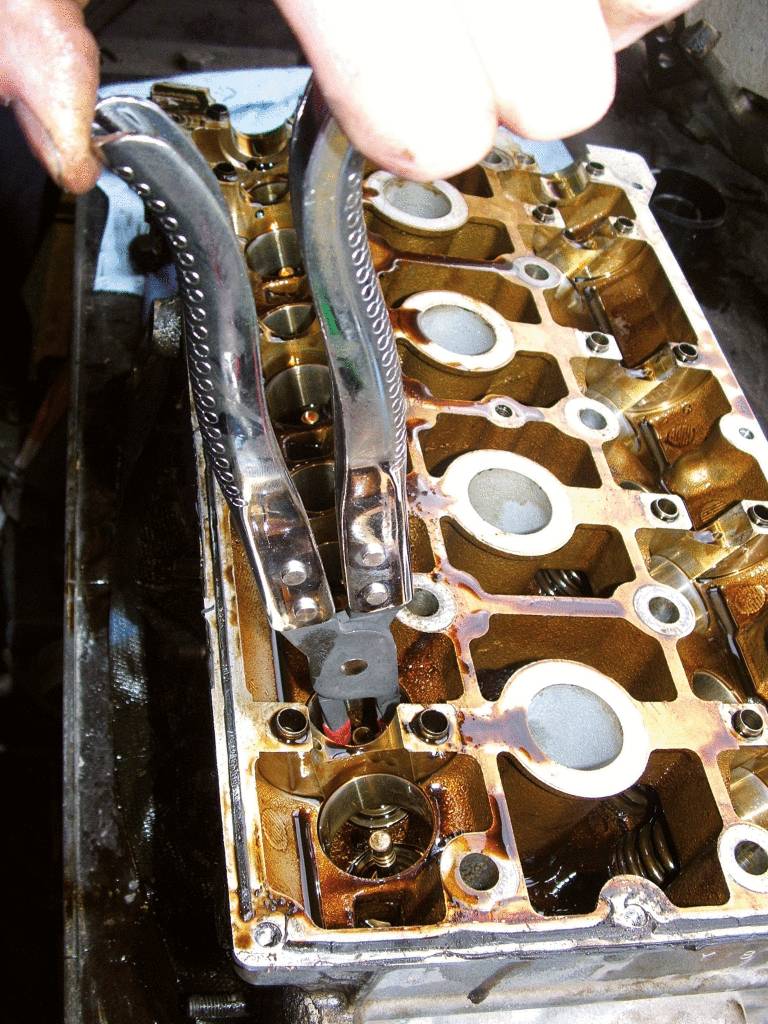 A person is working with pliers on an engine cylinder head, surrounded by metal parts and oil.