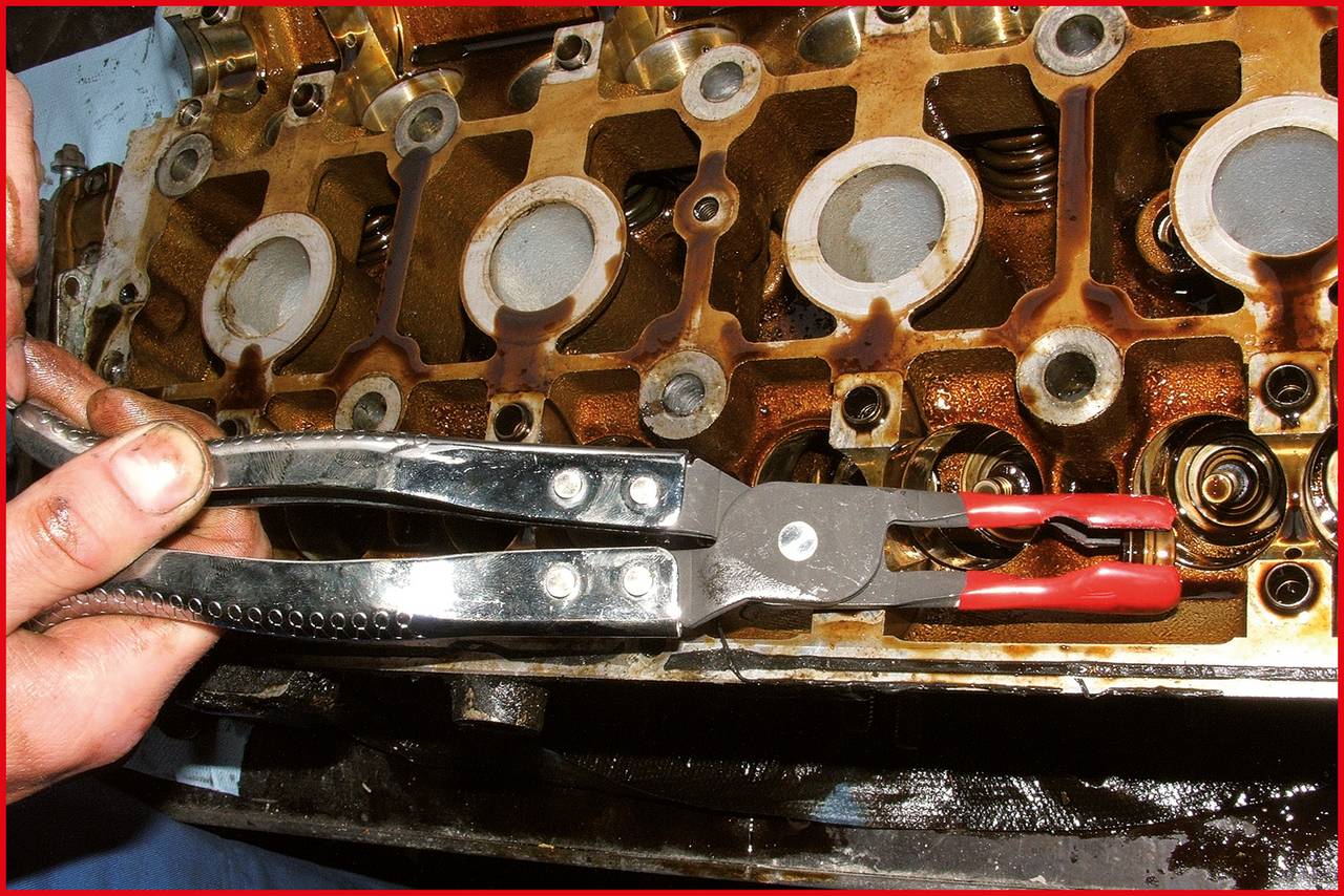 A mechanic holds pliers over a dismantled engine cylinder head. Pistons and valves are visible, work is being carried out.