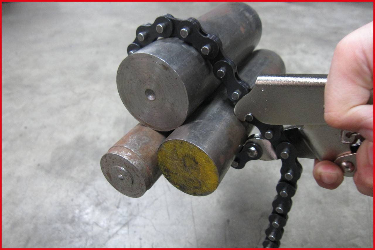 A chain tongs grips three metal cylinders on a concrete floor. A hand holds the tool, which is suitable for grasping cylindrical objects.