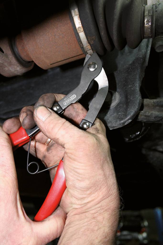 A person is repairing a car, using pliers to adjust a metal clip on a thick rubber hose or pipe. Their hands are dirty.