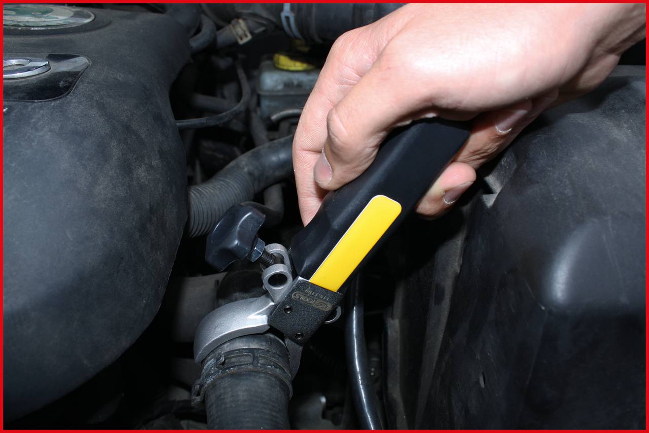A hand reaches for a black tool in the engine bay of a vehicle. Visible cables and metal components surround the tool.