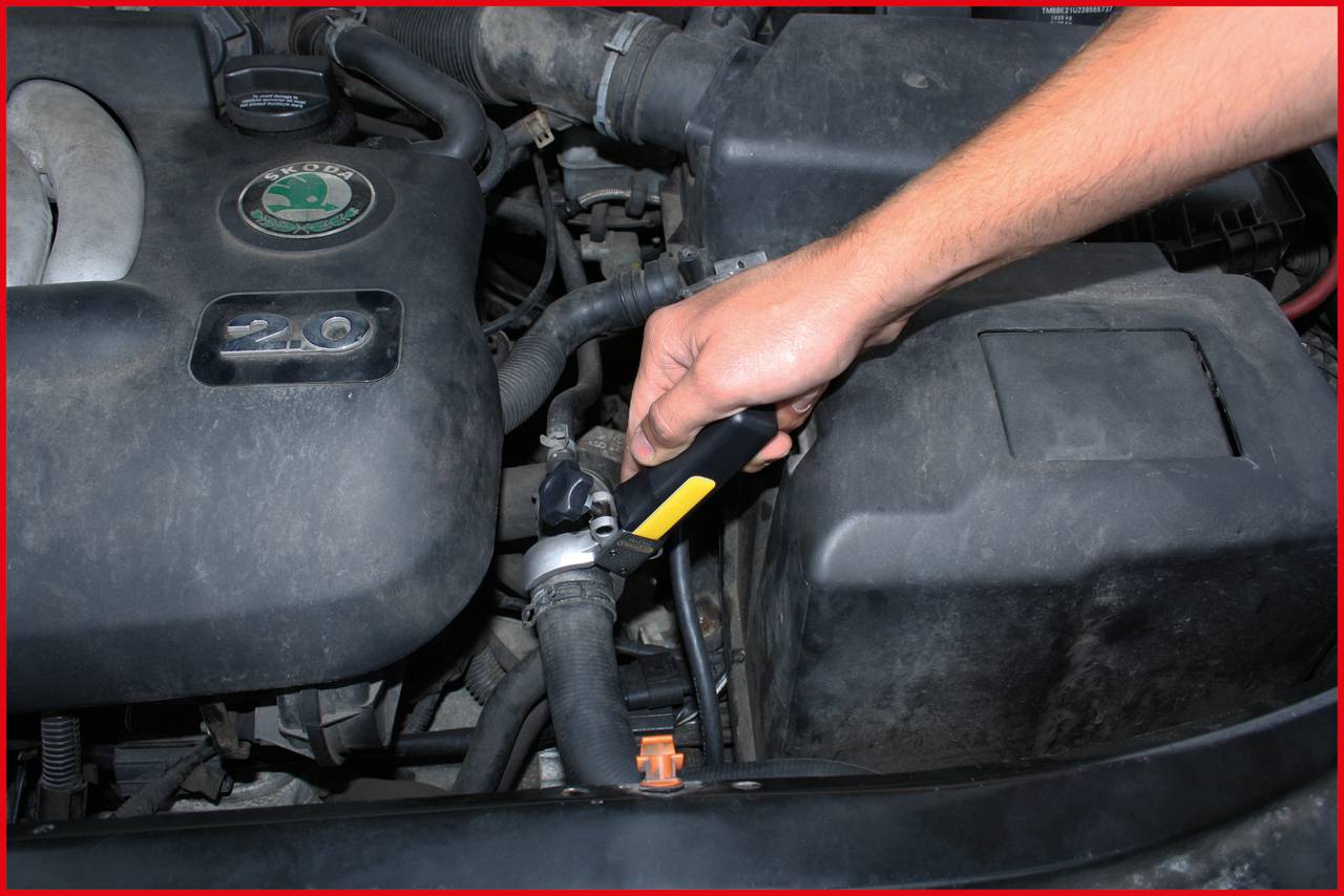 A person is checking the radiator hose of a car by applying a tool. Engine compartment with visible parts in the background.