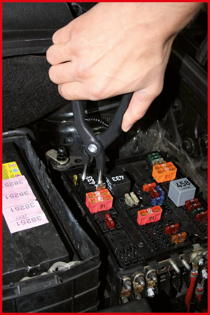 A hand is using pliers to remove a fuse in a car fuse box. The box is next to a car battery.