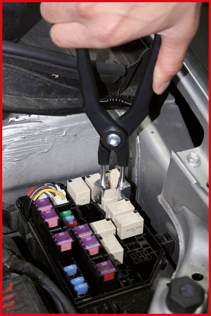 A hand is holding pliers and removing a fuse from a vehicle's fuse box, surrounded by several coloured fuses.