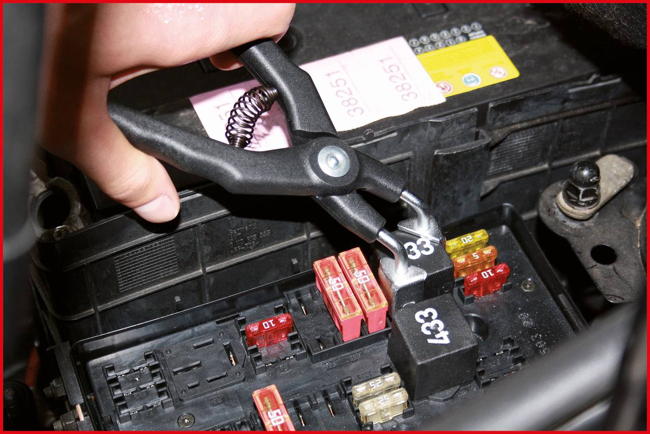 A hand is holding a clamp on a car fuse. Various coloured fuses and relays can be seen in the fuse box.