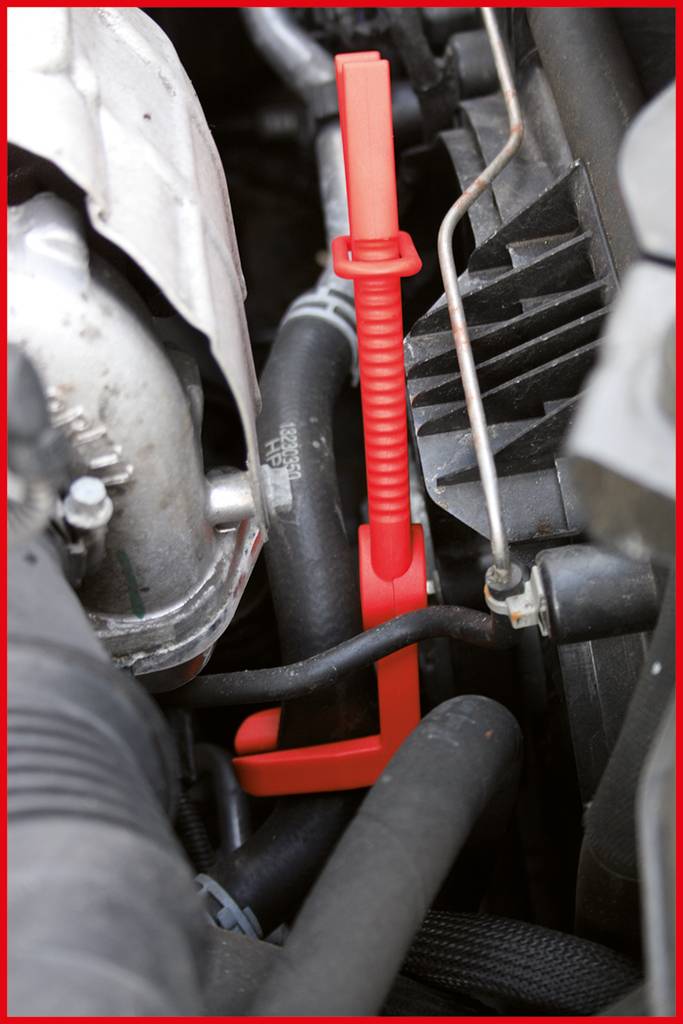 Red oil dipstick in an engine compartment; near the coolant hoses and other engine components. Essential for checking the engine's oil level.