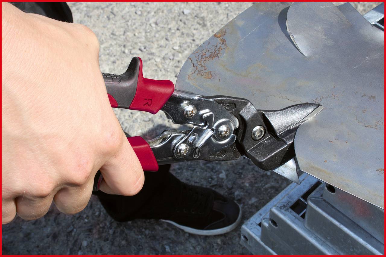 A hand holds a red metal shears cutting a piece of sheet metal. The shears point towards an edge of the metal, with the background out of focus.