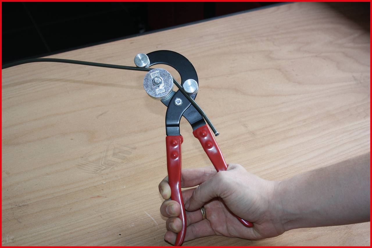 A hand is holding pliers that are bending a metal band. The pliers have red handles and are being used on a light-coloured wooden table.