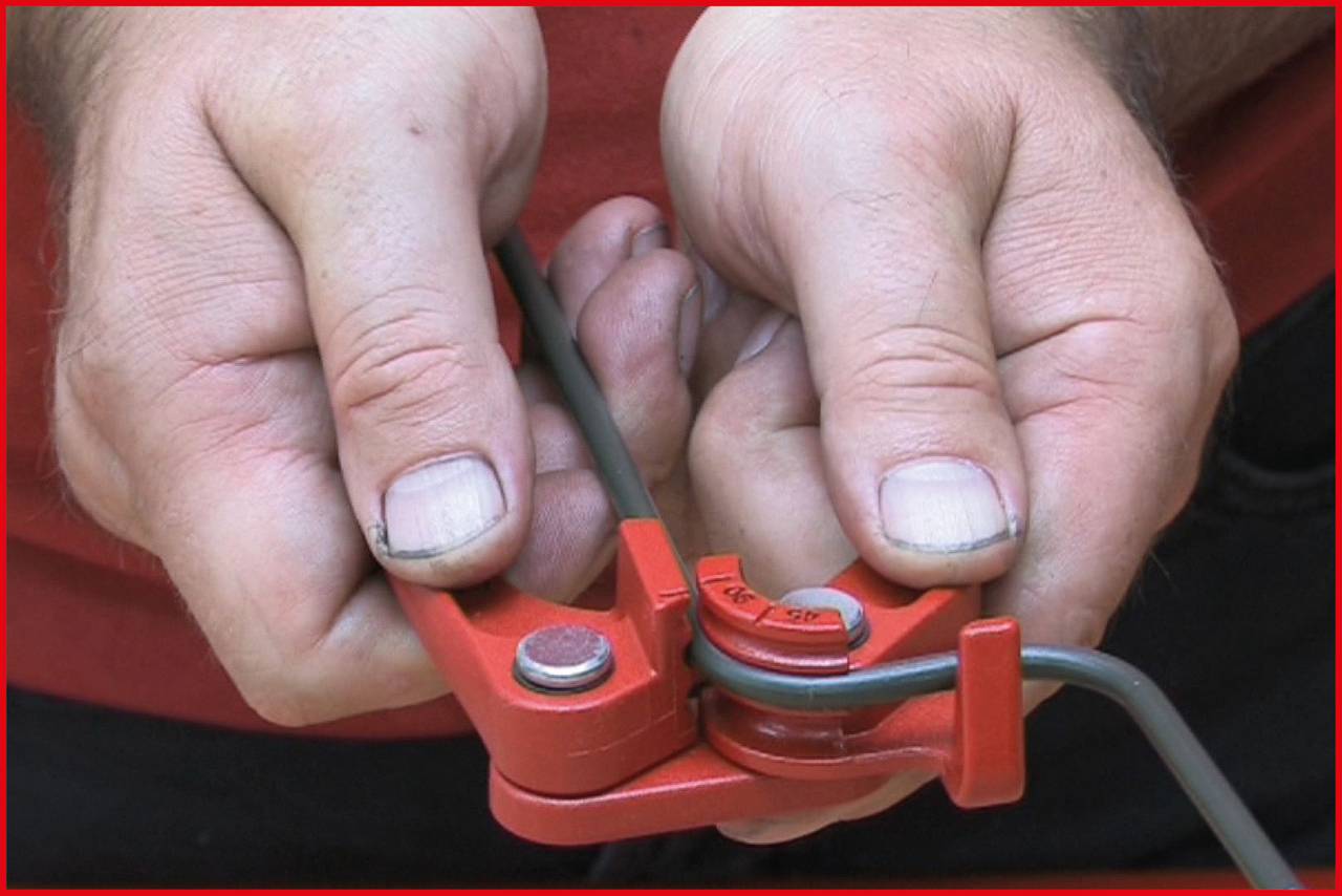 A person is bending a metal pipe with a red pipe bender. Hands focused on the workpiece.