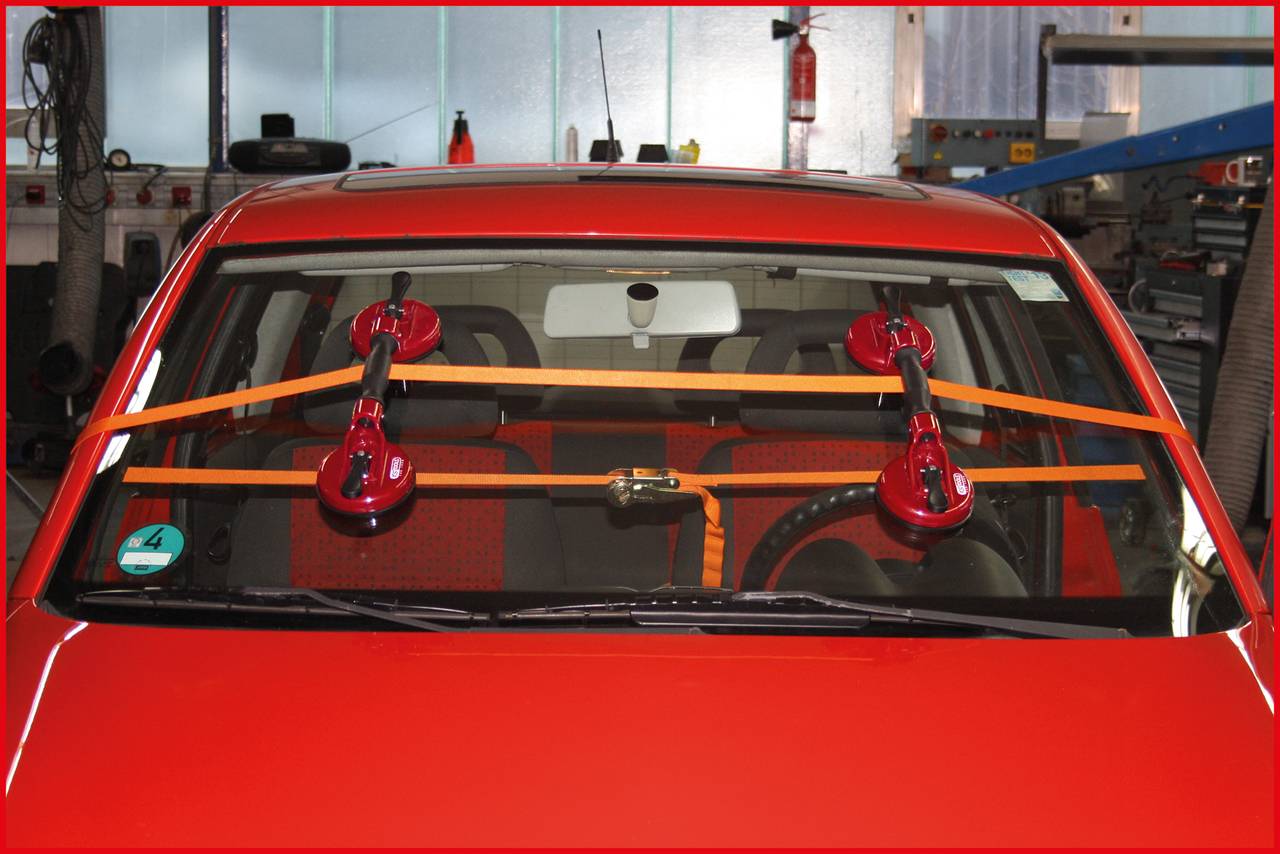 A red car with a secured windscreen fastened with suction cups and straps is parked in a workshop.