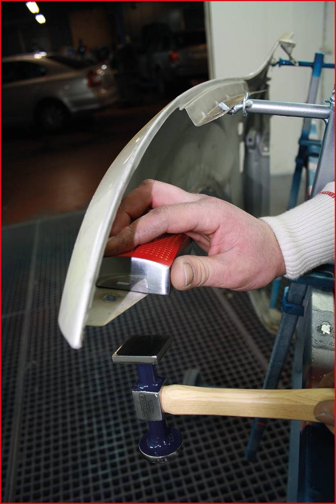 A person is repairing a car bumper with a tool for removing dents. Cars can be seen in the background of a workshop.