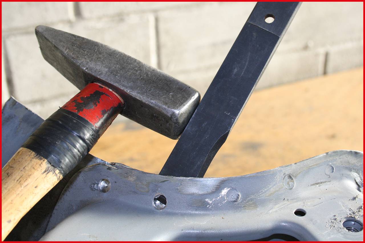 A hammer is leaning against a piece of metal resting on a table. A brick wall is visible in the background.