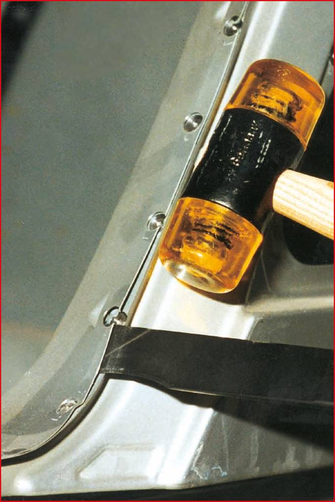 A hammer strikes a car body panel near a row of rivets. The hammer has a yellow rubber head and a wooden handle.