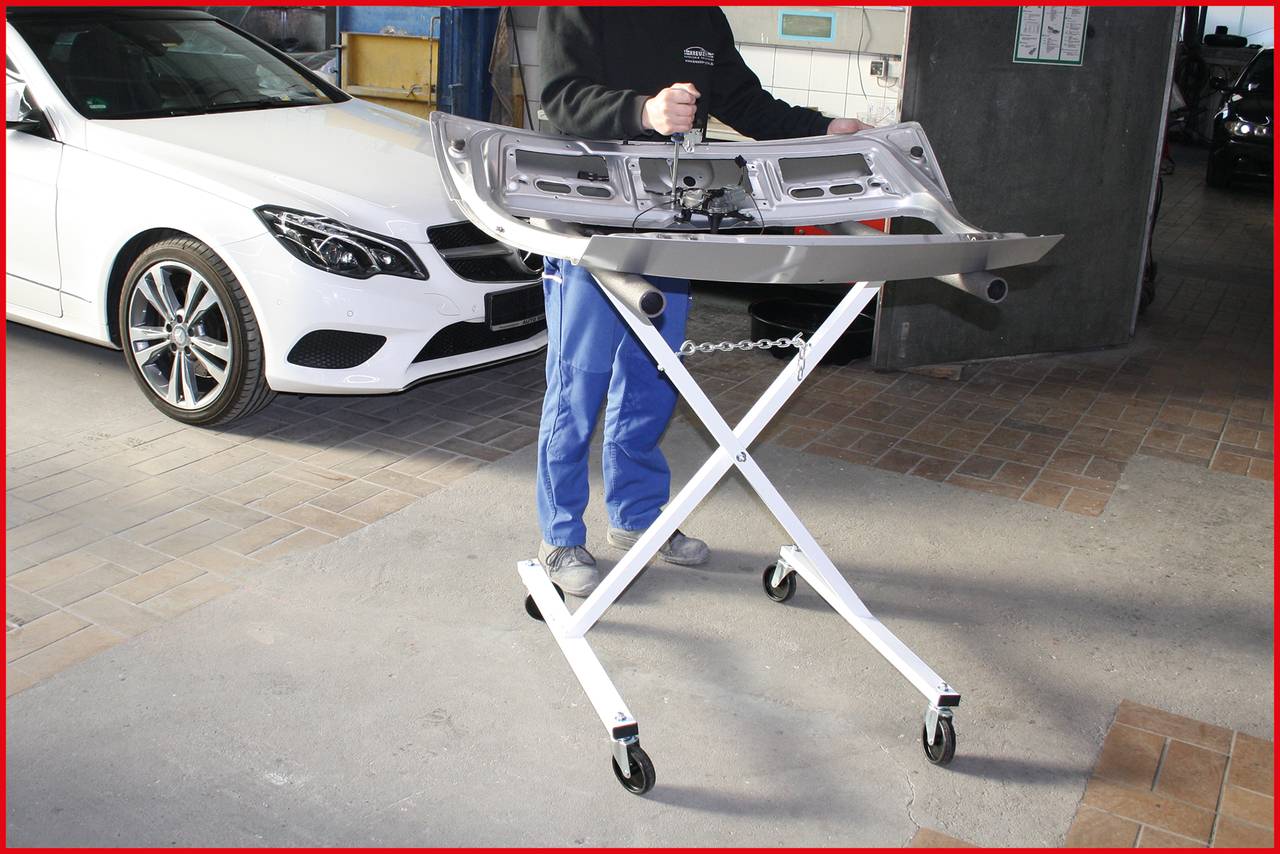 A person is working with an automotive part on a mobile trolley in a workshop. A white car is standing in the background.