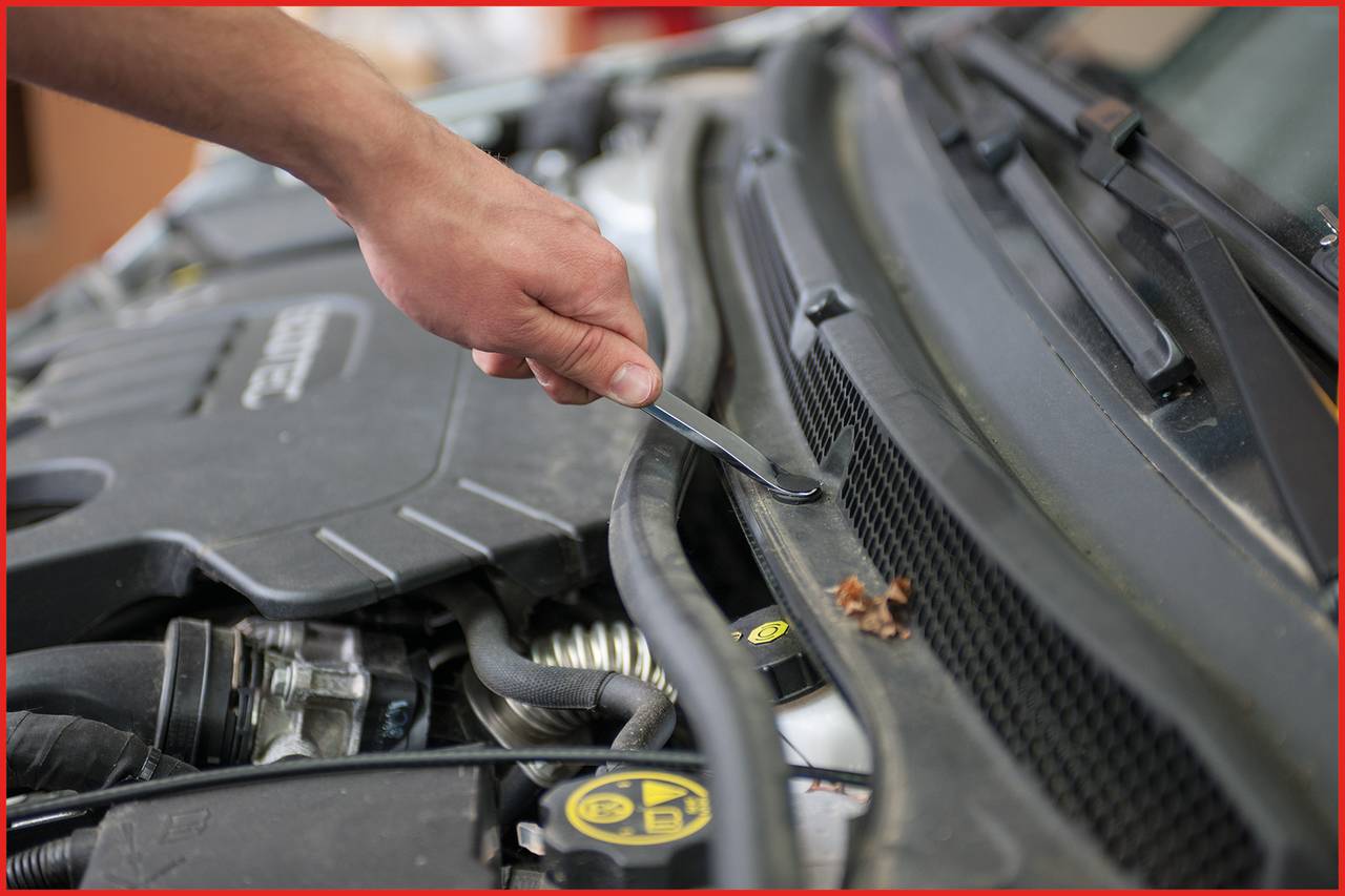 A hand opens the bonnet of a car with a screwdriver. Mechanical parts and the brand name are partially visible.