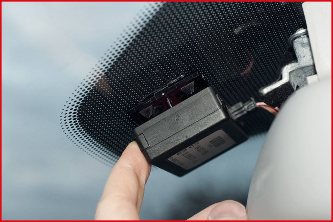 'A finger points to a black, rectangular device fixed behind a car's rear-view mirror on the windscreen.'